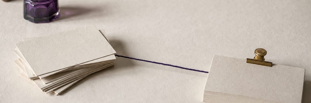 Two small stacks of blank index cards on pale paper, the left one loose and fanned, the right squared flush under a brass clip, a taut violet thread between them.